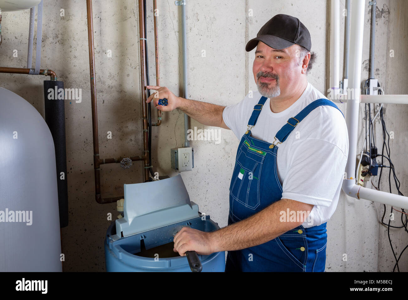 Freundlich lächelnden Workman in Latzhosen installieren oder die Arbeit an einem Wasserenthärter in einem Hauswirtschaftsraum drehen zu lächeln in die Kamera Stockfoto