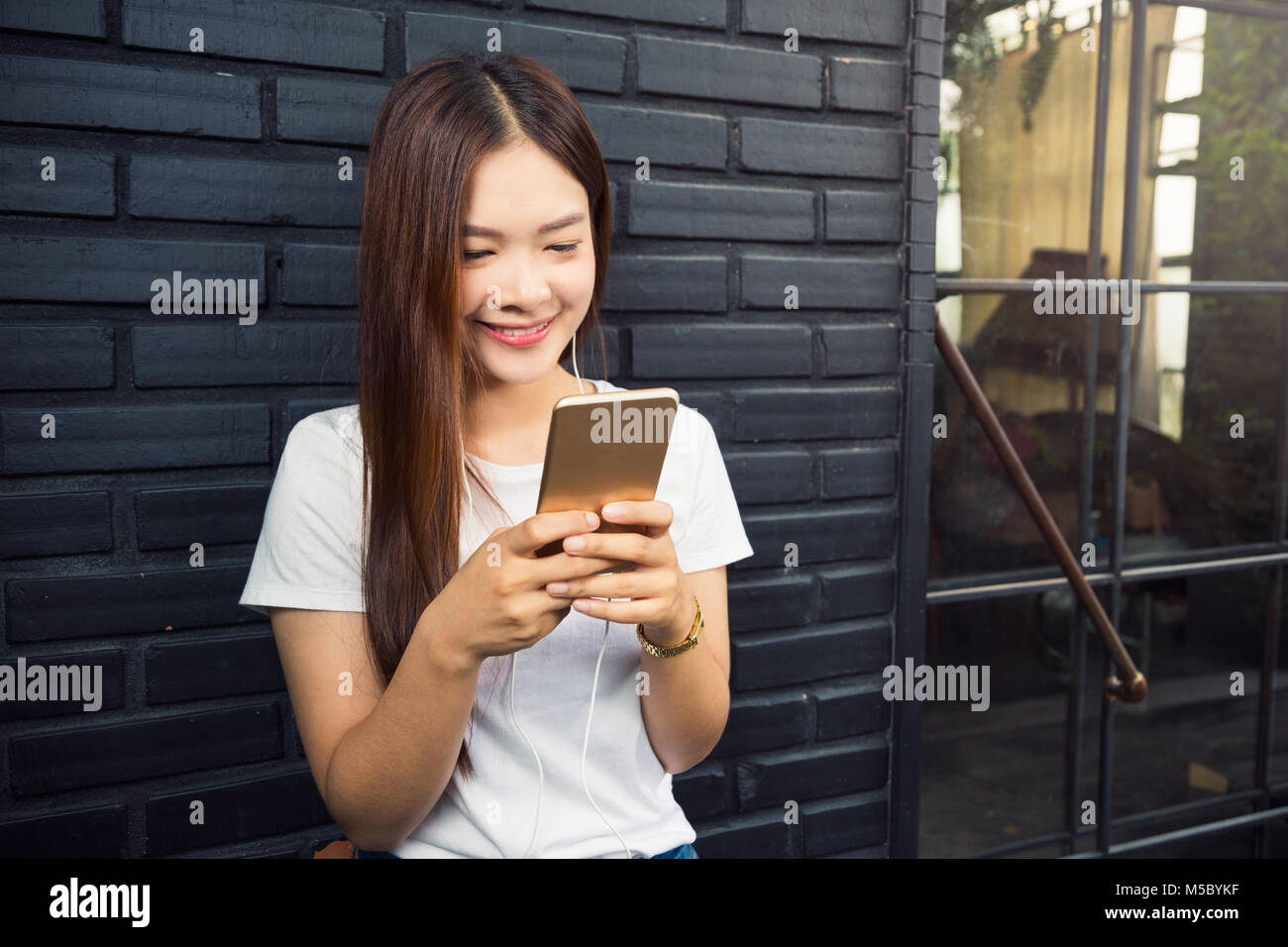 Junge schöne asiatische Frau, gegen dunkle Mauer mit Ihrem Smart Phone zu hören, online Musik ansehen, und mit ihren Freunden, gute Stockfoto