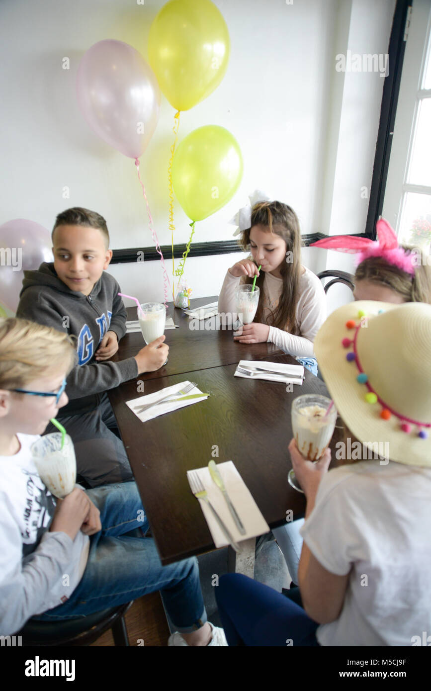 Fünf junge Kinder sitzen auf einer Party Tisch essen gebratene Nahrung und trinken Milchshakes - es gibt Ballons und Party Dekorationen Stockfoto