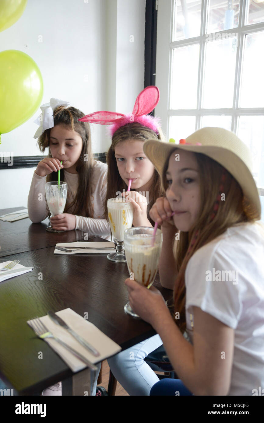 Drei junge Kinder sitzen auf einer Party Tisch essen gebratene Nahrung und trinken Milchshakes - es gibt Ballons und Party Dekorationen Stockfoto