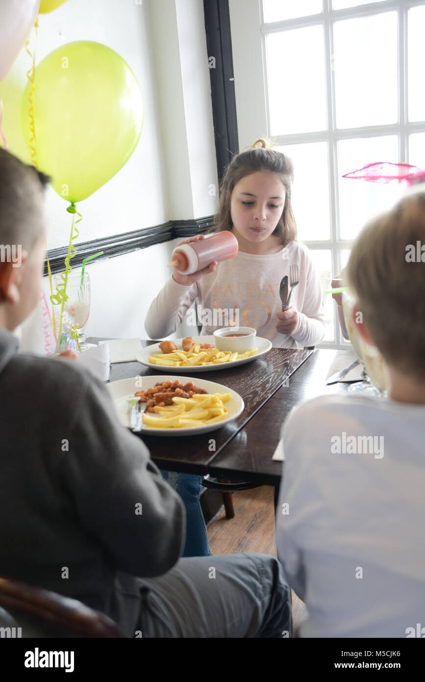 Drei junge Kinder sitzen auf einer Party Tisch essen gebratene Nahrung und trinken Milchshakes - es gibt Ballons und Party Dekorationen Stockfoto