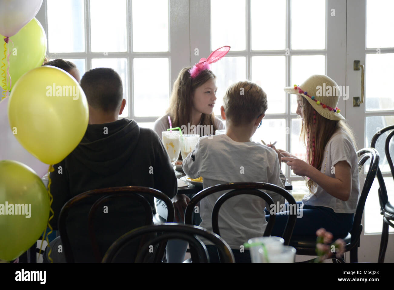 Vier junge Kinder sitzen auf einer Party Tisch essen gebratene Nahrung und trinken Milchshakes - es gibt Ballons und Party Dekorationen Stockfoto