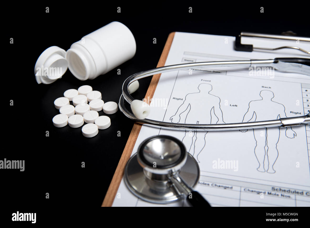 Weiße Pillen und eine weiße Flasche, zusammen mit einem Stethoskop und ein medizinisches Diagramm, werden über einen schwarzen Hintergrund. Stockfoto