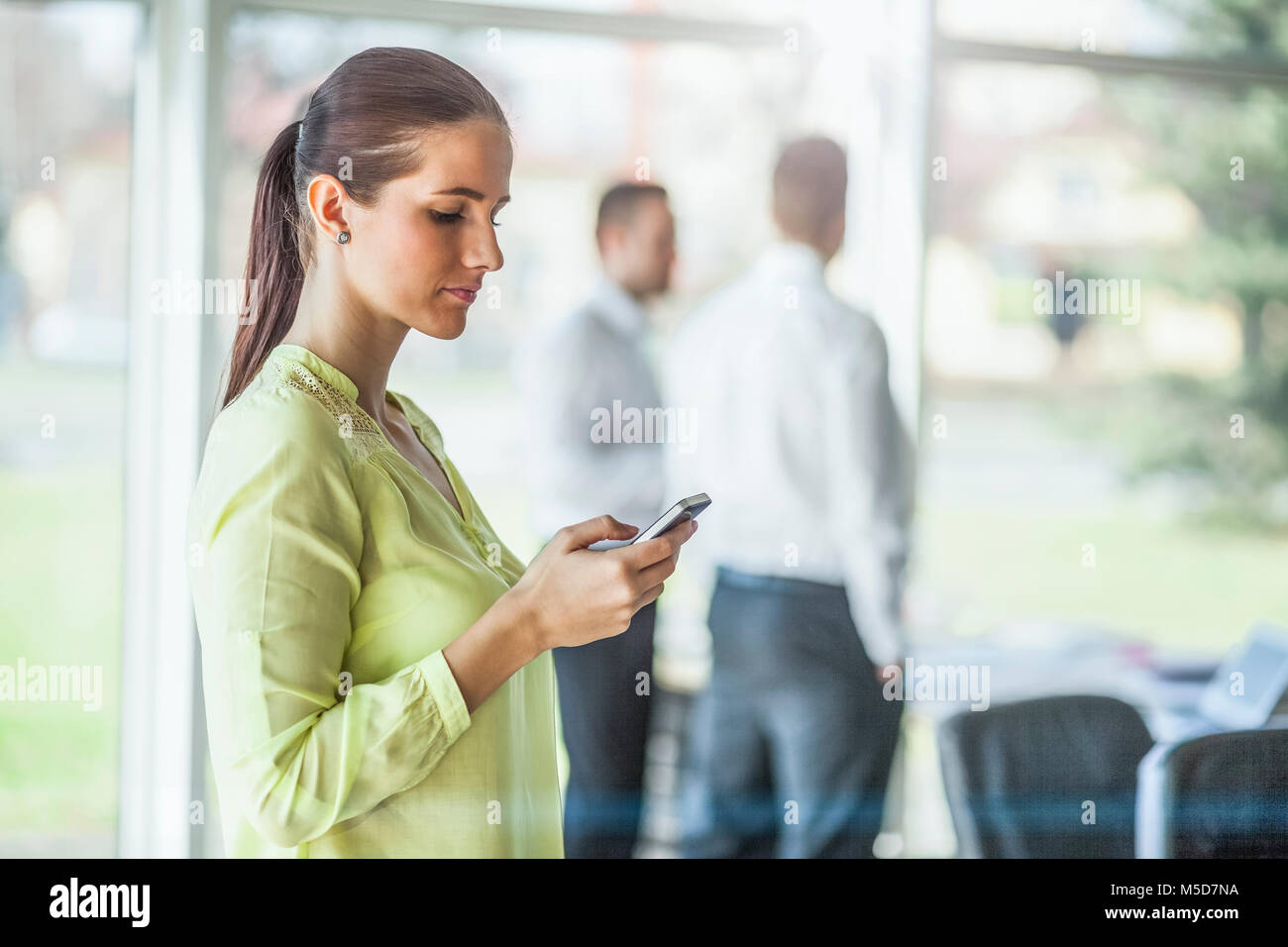 Seitenansicht der jungen Geschäftsfrau mit Handy mit männlichen Kollegen im Hintergrund im Büro Stockfoto