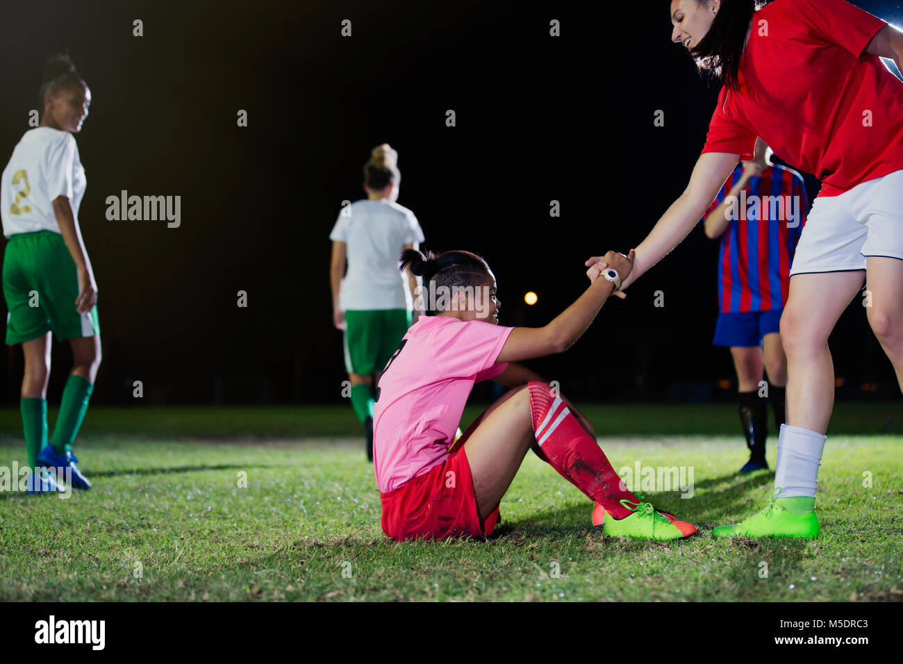 Junge weibliche Fußball-Spieler helfen gefallenen Teamkollegen auf Feld bei Nacht Stockfoto