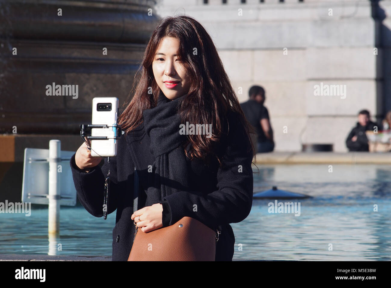 Eine junge Frau, die eine orientalische selfie im Winter Sonne neben einem Pool in Trafalgar Square in London mit einem selfie Stick Stockfoto