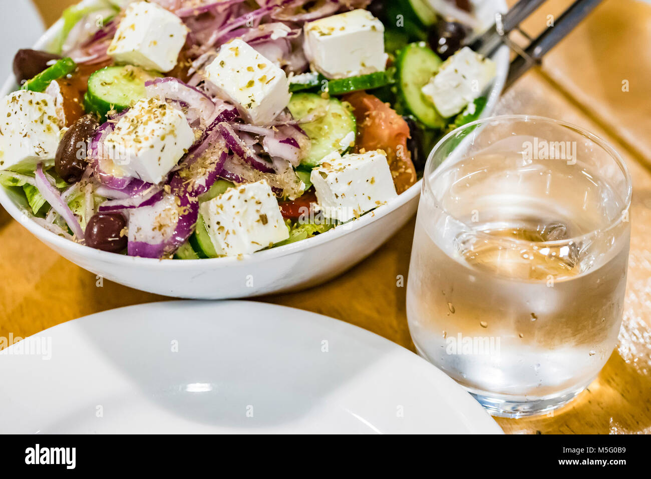 Frischer Salat, Teller, Glas Wasser auf dem Tisch, Nahaufnahme. Griechischer Salat mit Feta Käse, gesundes Essen Konzept. Stockfoto