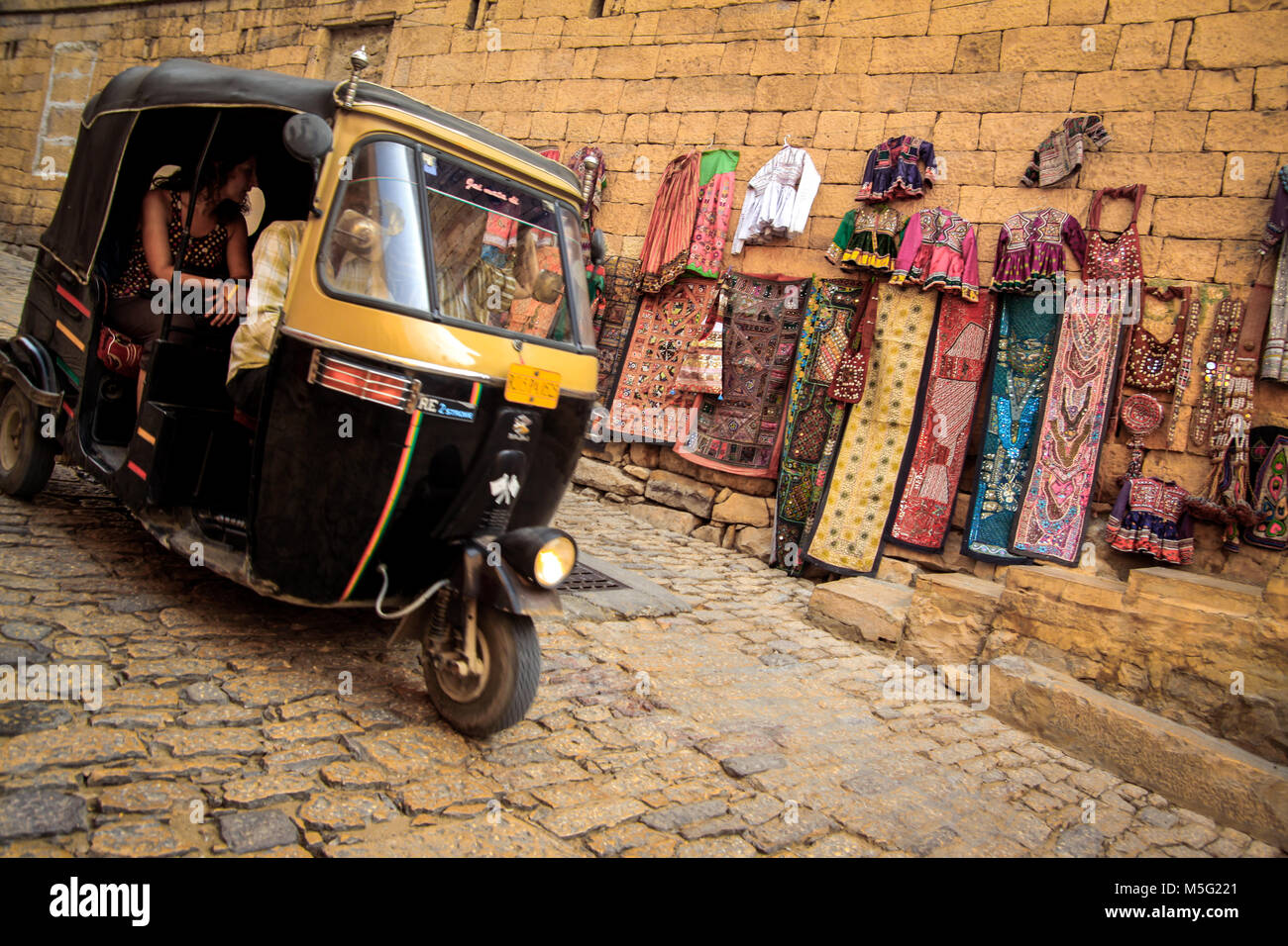 Indische Auto-rikscha Touristen transportieren, das in der ummauerten Heritage Area von Jaisalmer in Rajasthan. Bunte Rajasthani Kleidung hängen von der Wände Stockfoto