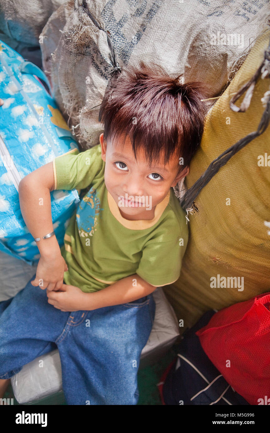 Eine Vorschule philippinischen Jungen mit großen, faszinierenden asiatischen Augen und ein leichtes Lächeln sitzt unter cargo Taschen auf einer Fähre in den Philippinen. Stockfoto