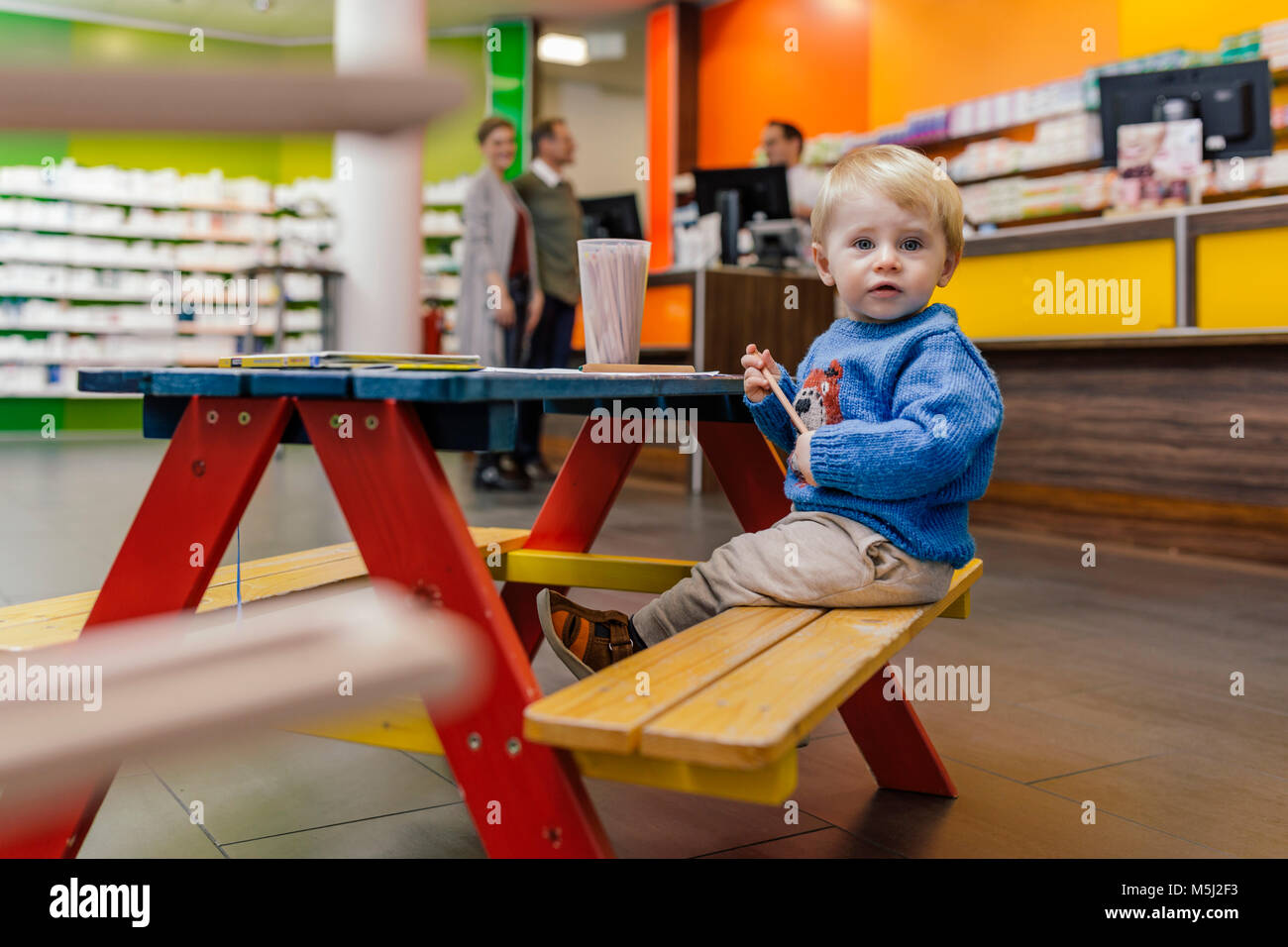 Kleiner Junge sitzt auf der Bank in der Apotheke mit Eltern im Hintergrund Stockfoto