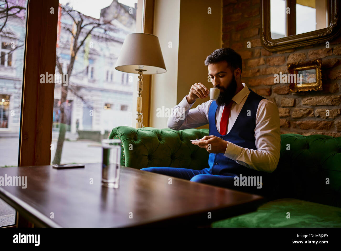 Modische junge Mann sitzt auf der Couch in einem Cafe, Kaffee zu trinken und mit Handy Stockfoto