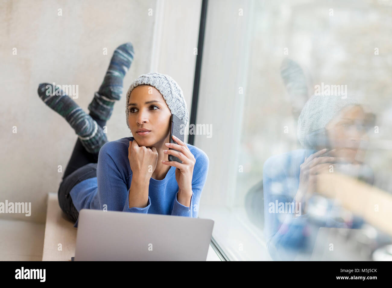 Porträt der jungen Frau am Telefon liegend mit Laptop auf Fensterbank Stockfoto