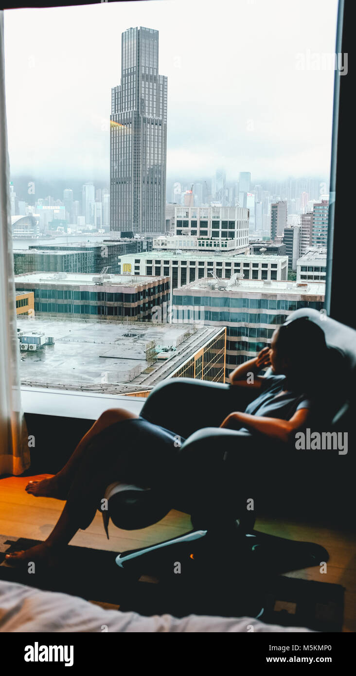 Eine Frau touristische schaut aus Ihrem Hotelzimmer im Hotel Icon an der Skyline von Kowloon, Hong Kong an einem bewölkten Tag Stockfoto