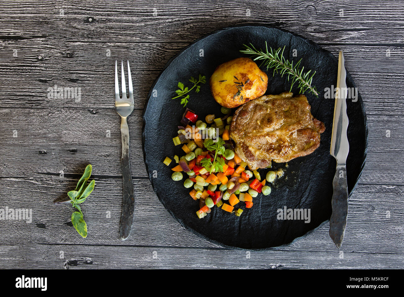 Schweinefleisch vom Grill mit gebratenem Gemüse und gebackene Kartoffel auf einer schwarzen Platte Stockfoto