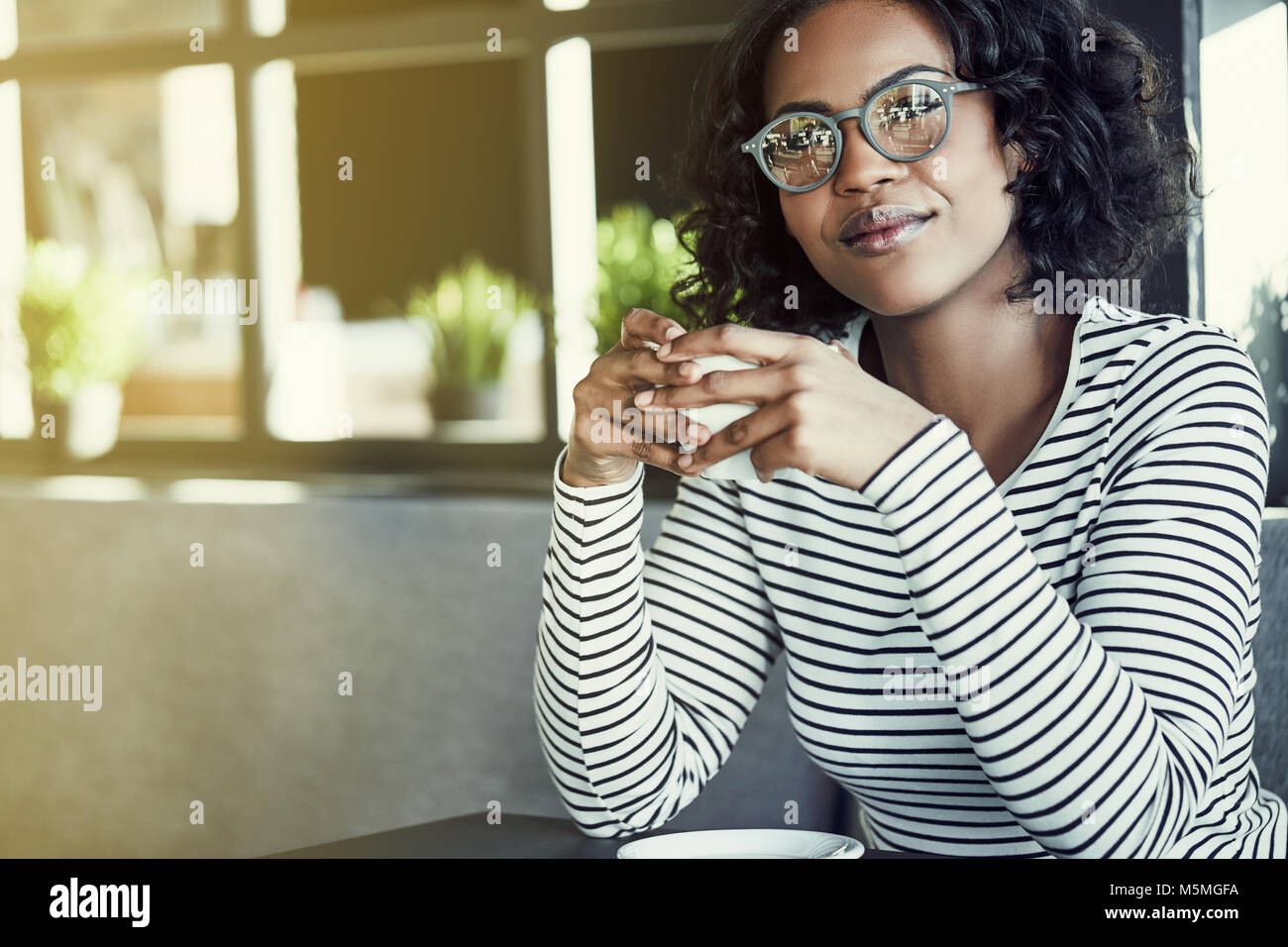 Lächelnden jungen afrikanischen Frau Brille trägt allein sitzen an einem Tisch in einem Cafe Trinken einer Tasse Kaffee Stockfoto