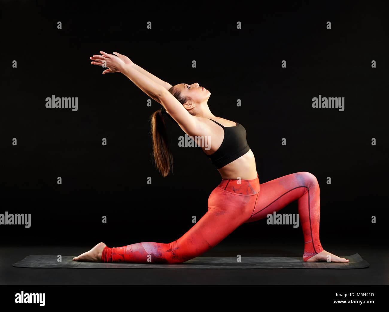 Passende Frau zeigt eine niedrige Ausfallschritt pose in Yoga, Stretching und Bogenschiessen zurück Ton ihre Muskeln, Seitenansicht über einen schwarzen Hintergrund zu Stockfoto