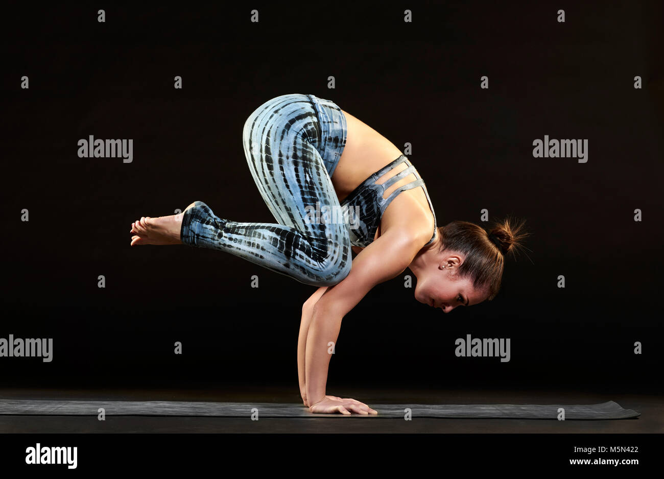Athletische Passform junge Frau, die eine Krähe im Yoga Balancieren auf ihre Arme mit Ihrem Körper in eine gewellte, seitliche Blick auf Schwarz Stockfoto
