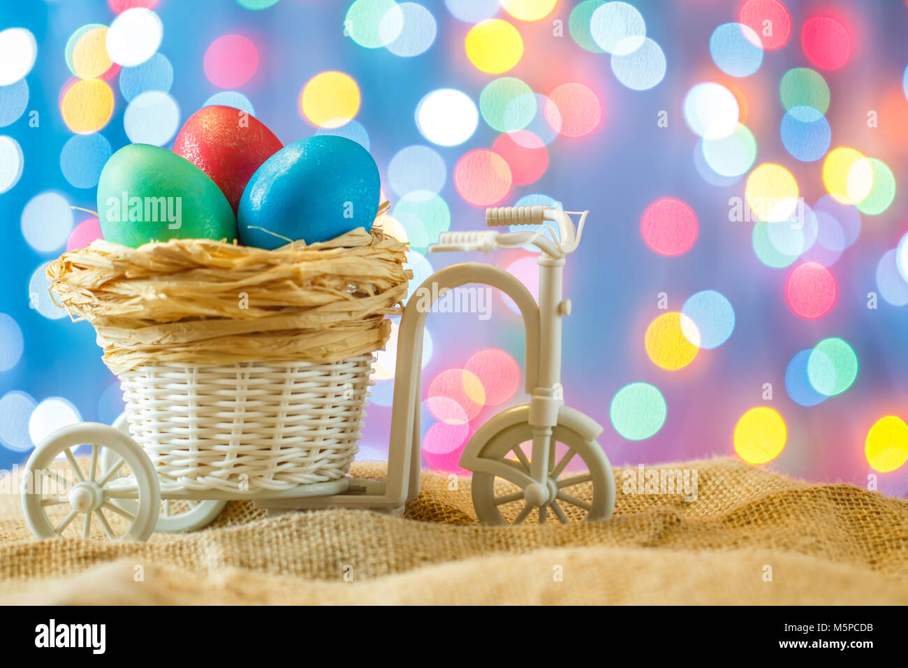 Ostern Karte, farbige Eier im Nest. Spielzeug Fahrrad mit einer Karre. Frohe Ostern. Blau bokeh Hintergrund. Spielzeug Fahrrad mit Warenkorb Stockfoto
