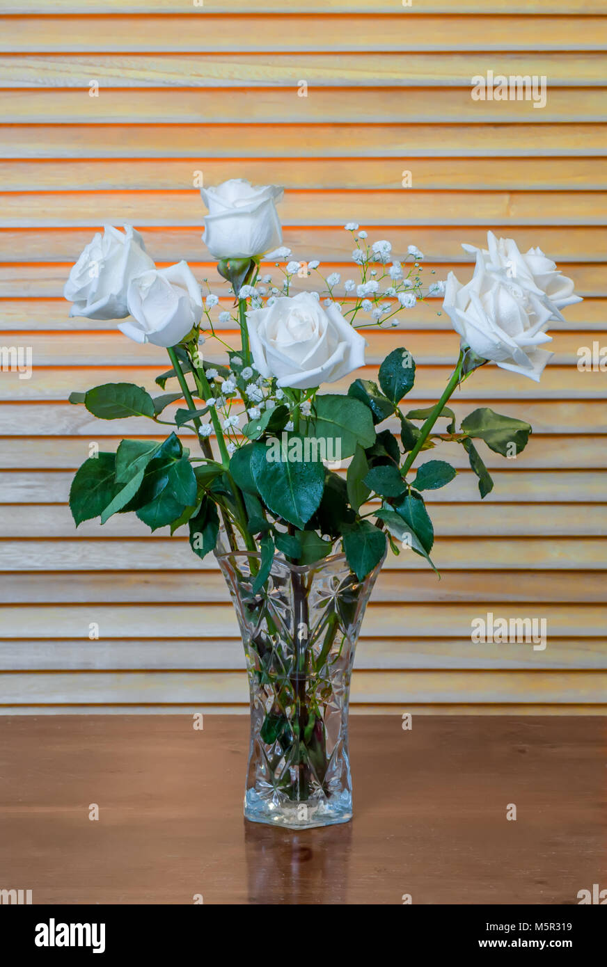 Die Anlage ist ein Blumenstrauß aus den weißen Rosen mit grünen Blättern und kleinen, weißen Blüten, in ein Glas Vase auf einem hölzernen braunen Tisch, vor dem Hintergrund der Ihr Stockfoto