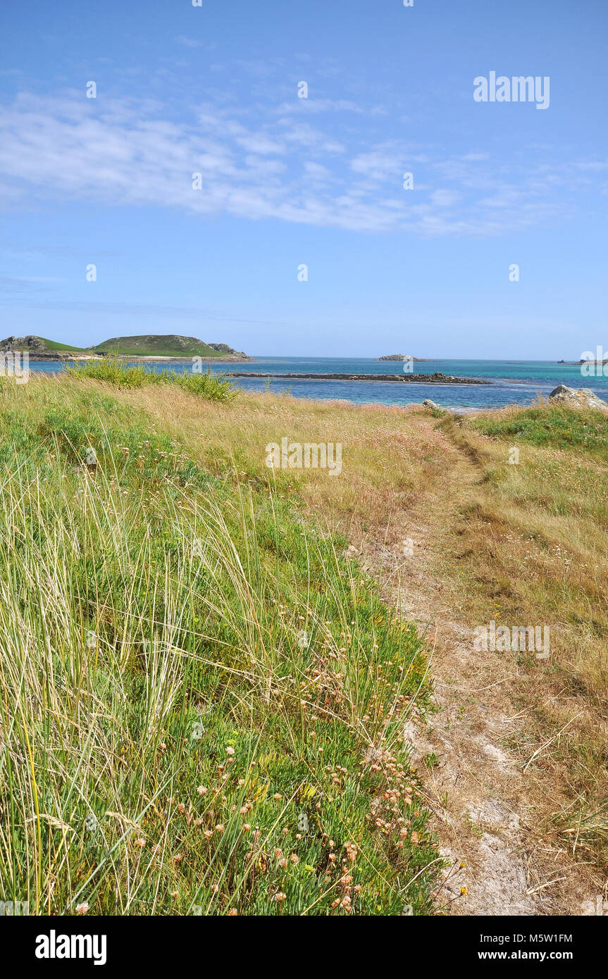 Sonnige Strände der Scilly Isles, Großbritannien. Blick auf eine Bucht mit Gras im Vordergrund und blauem Himmel mit Wolken. Stockfoto