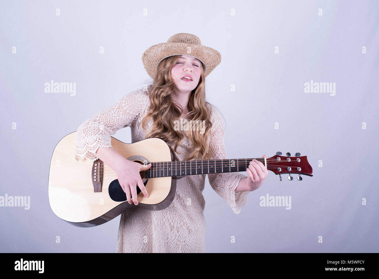 Der 12-jährige Mädchen mit langen, schmutzig blonden Haar, trug Stroh Hut singen mit geschlossenen Augen, spielen Akustische 6-saitige Gitarre, weißer Hintergrund Stockfoto