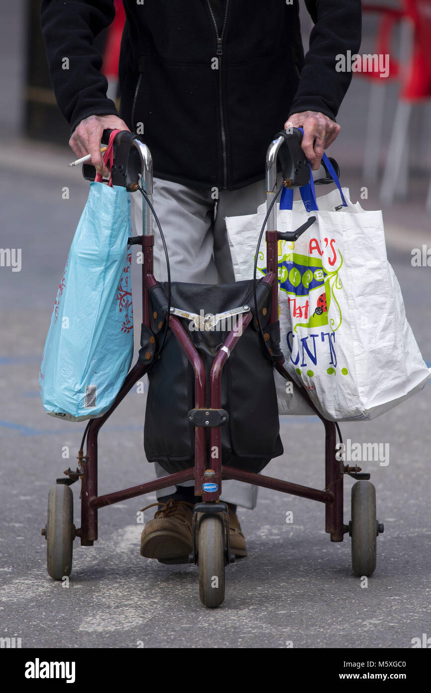 Ein alter Rentner mit einer Gehhilfe für Unterstützung, während auf der Straße tragen Einkaufstaschen in Cardiff, Wales, UK. Stockfoto
