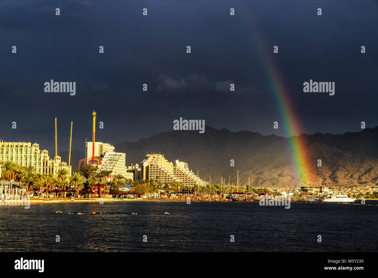 Rainbow und dunkle Regenwolken über aufgehellt Strand und Hotels in Eilat, Israel. Golf von Aqaba am Roten Meer. Schwimmer Training für nächsten Tag Ironman Rennen Stockfoto