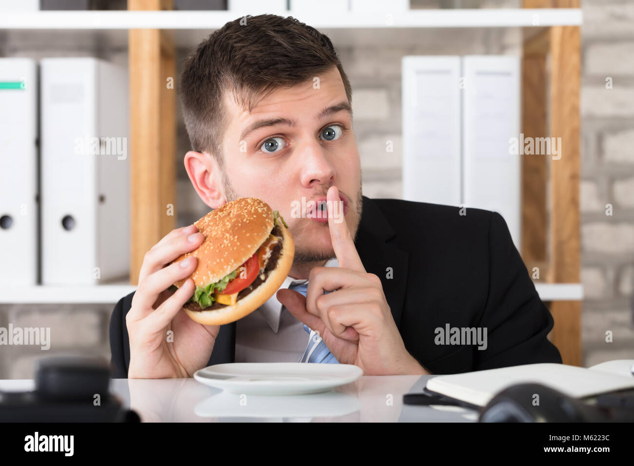 Porträt eines hungrigen Jungen Geschäftsmann Holding Burger mit Finger auf die Lippen im Büro Stockfoto