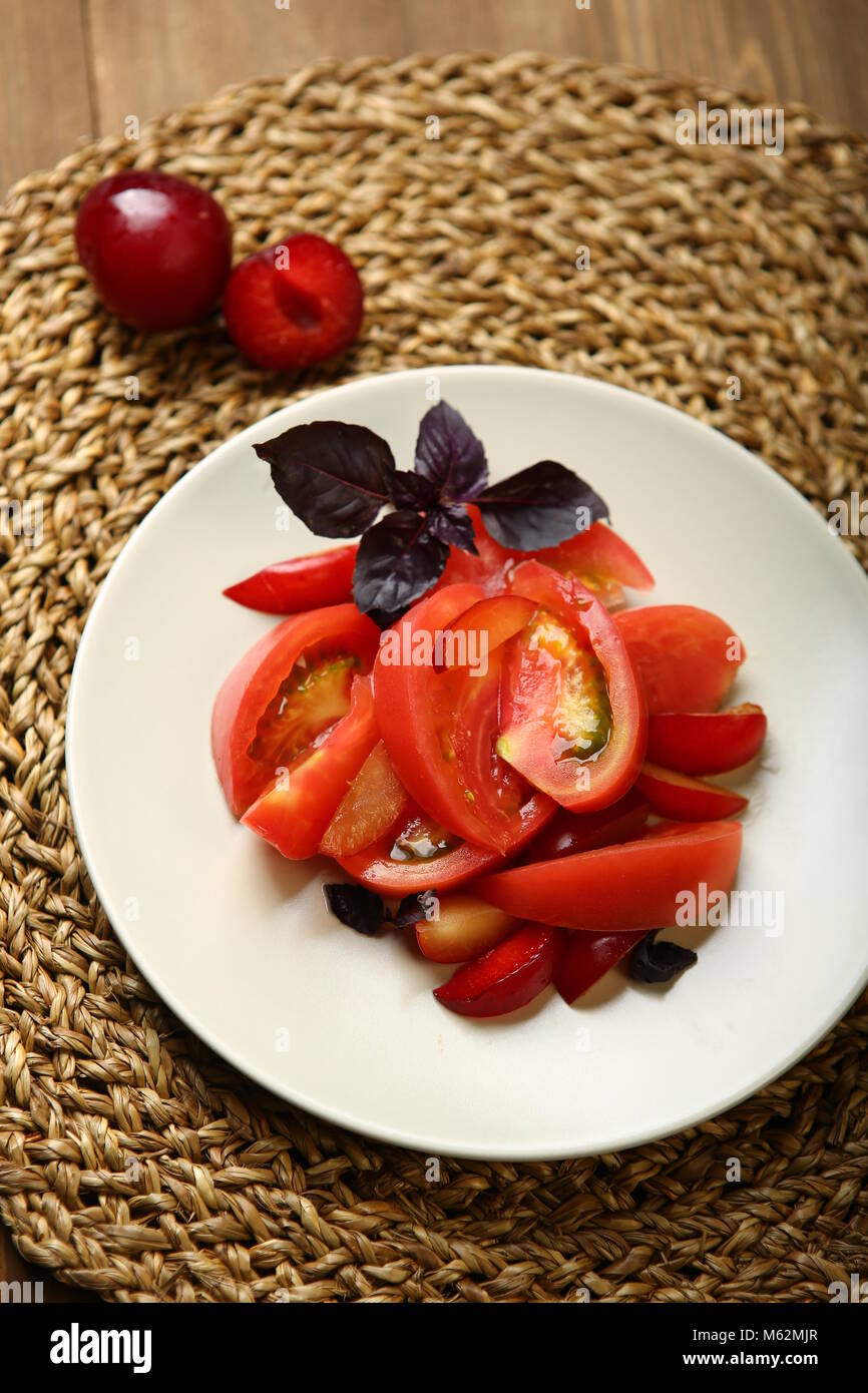Salat von Tomaten und rote Pflaumen mit Basilikum für eine gesunde Ernährung. Kalorienarme Nahrung für Körper Gewicht kontrollieren. Schnelle Snacks. Auf einem Holztisch, ein Korb stehen. Stockfoto
