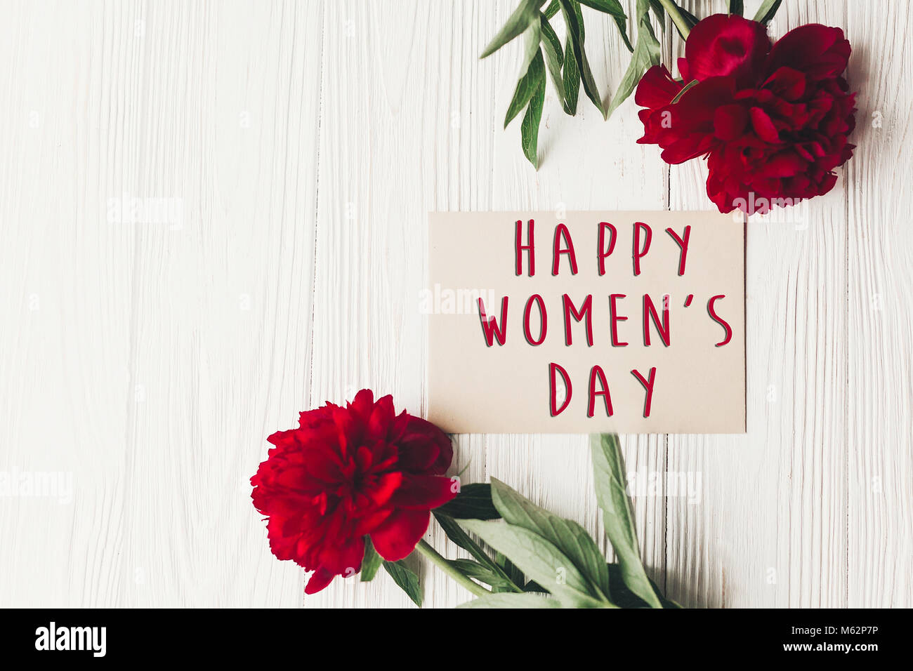 Der glückliche Frauen Tag Text Zeichen für das Handwerk Karte und schönen roten Pfingstrosen auf weißen Holzmöbeln rustikal Hintergrund, flach. Moderne Grußkarte. Tag der Frauen Stockfoto