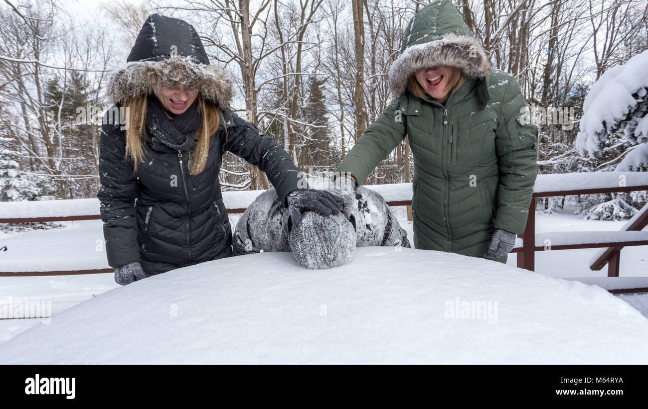 Zwei Yong Frauen im Winter Mäntel spielerisch zu drücken, um eine junge bemannt Gesicht in den Schnee Stockfoto