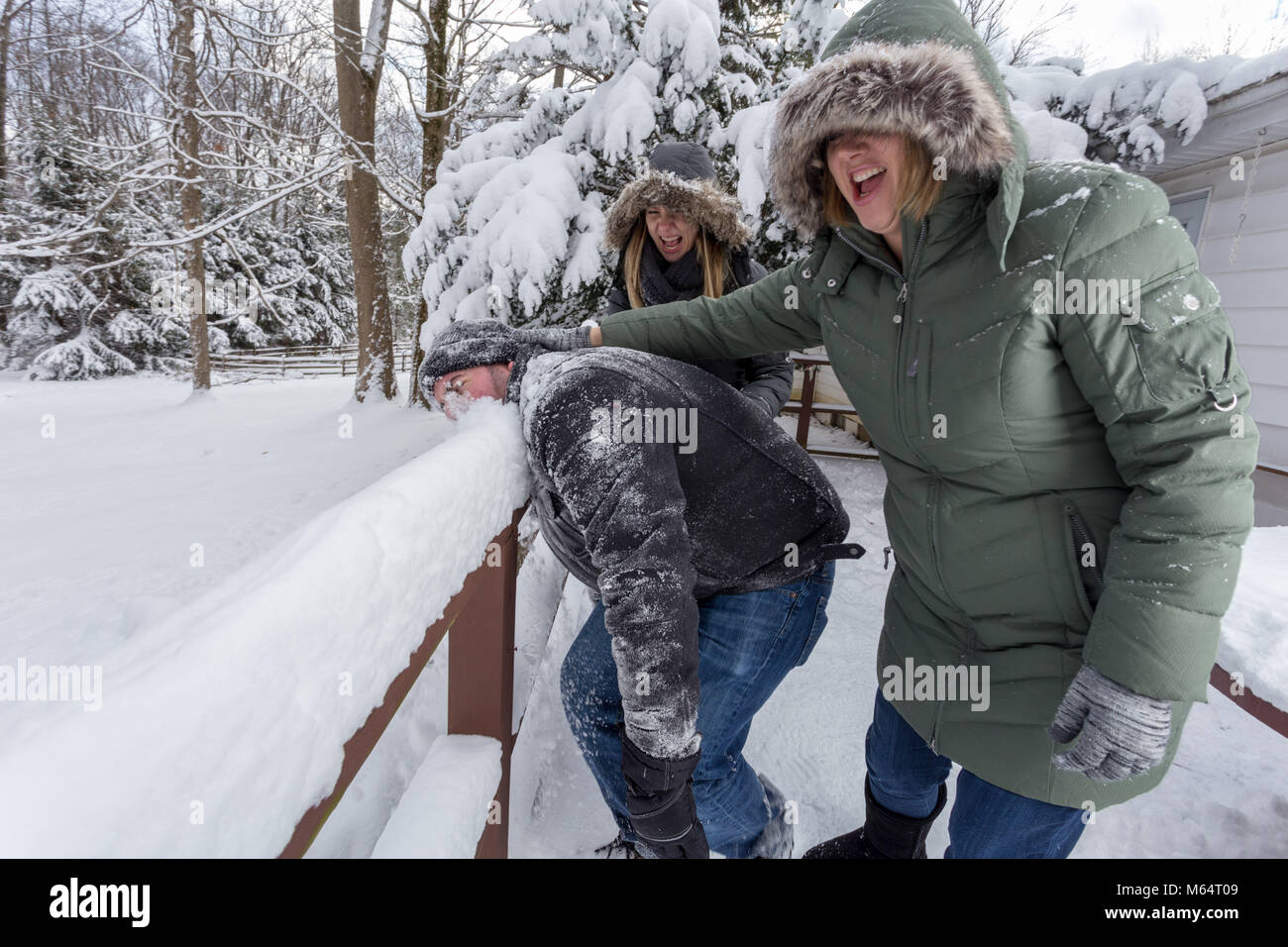 Zwei Yong Frauen im Winter Mäntel spielerisch zu drücken, um eine junge bemannt Gesicht in den Schnee Stockfoto