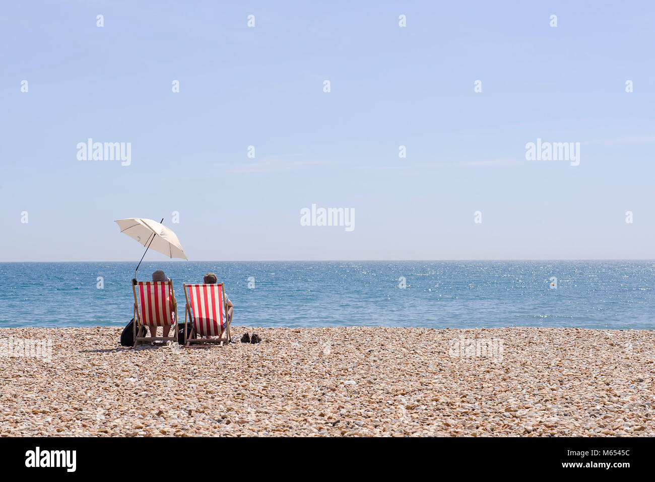 Die steigenden Temperaturen an der Südküste von England als Paar entspannen Sie sich auf traditionelle gestreifte Liegestühle mit Sonnenschirm auf einem glühend heißen Tag in Lyme Regis. Stockfoto