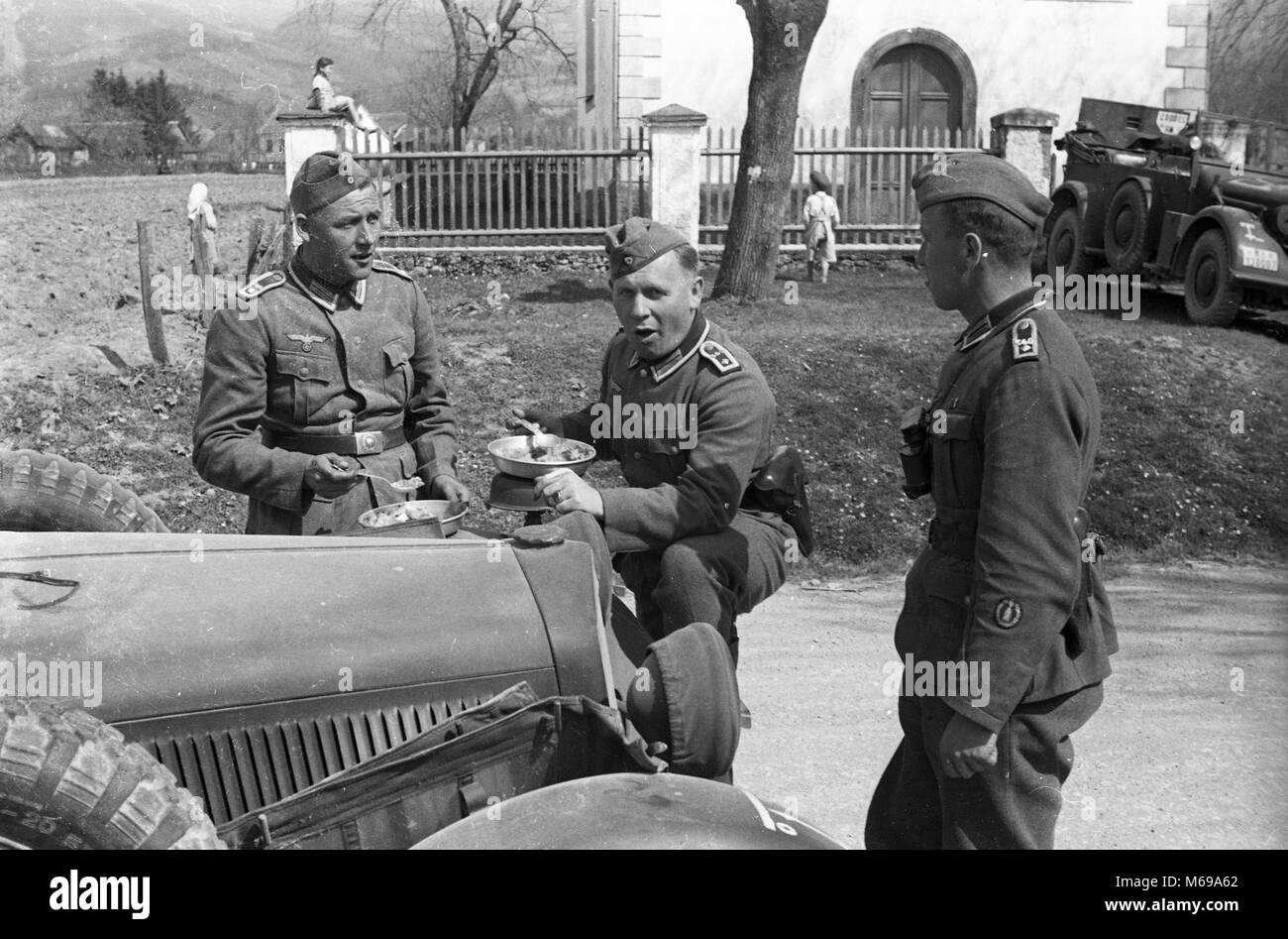 Deutsche Soldaten beim Mittagessen mit lokalen Kinder spielen im Hintergrund während der Invasion von Jugoslawien im April 1941. Stockfoto