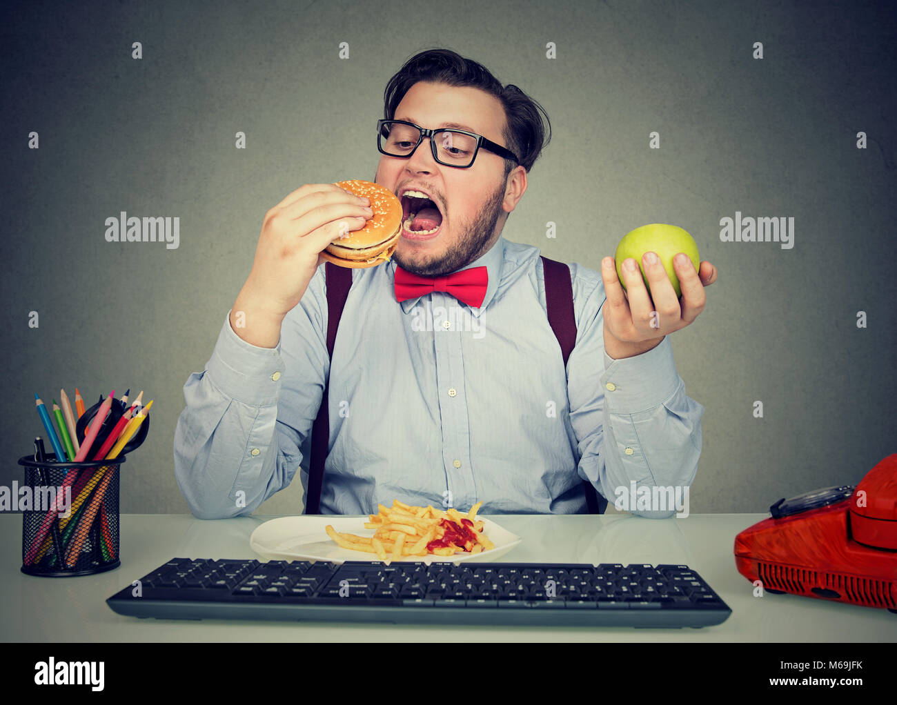 Junger Mann essen Hamburger mit Pommes anstelle von Apple in Bewegungsmangel im Büro. Stockfoto