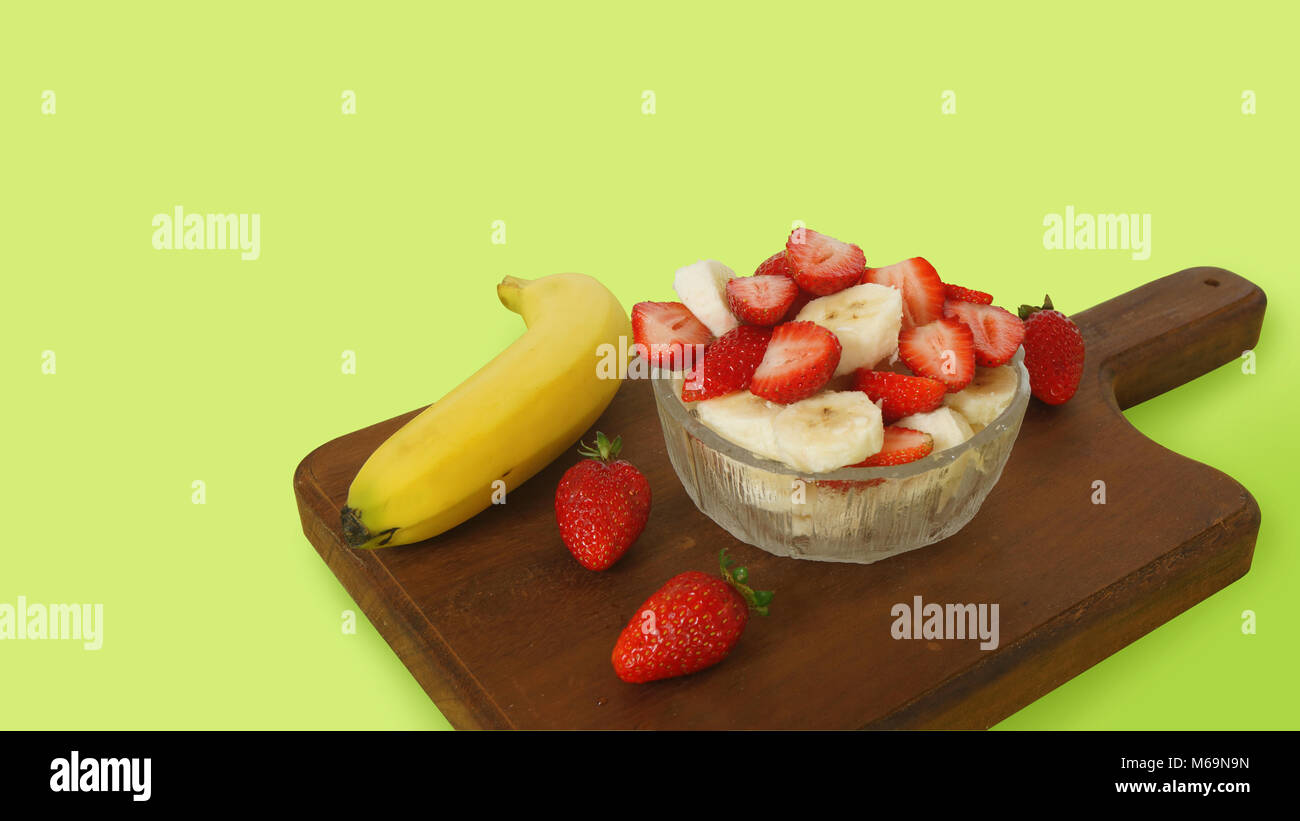 Glasplatte mit gehackt Erdbeeren und Bananen Stücke auf einem dunklen Holzplatte mit 3 Erdbeeren und eine Banane mit Schale auf einem grünen Zurück eingerichtet Stockfoto