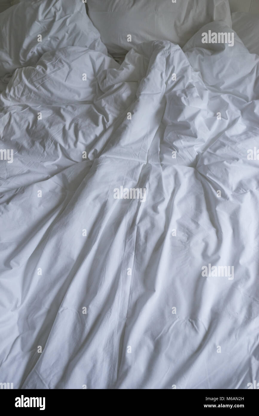 Ungemachten Bett mit einfacher weißer Bettwäsche in Nahaufnahme eines rumpled Bettdecke, Kissen und ein gepolstertes Kopfteil Stockfoto
