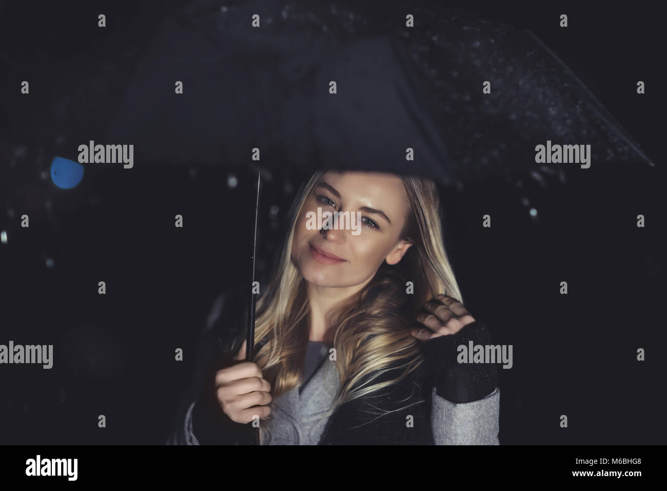 Glückliches Mädchen an regnerischen Nacht draußen, Porträt einer schönen blonden Frau mit Schirm im Regen, gute Laune in kaltem Wetter Stockfoto