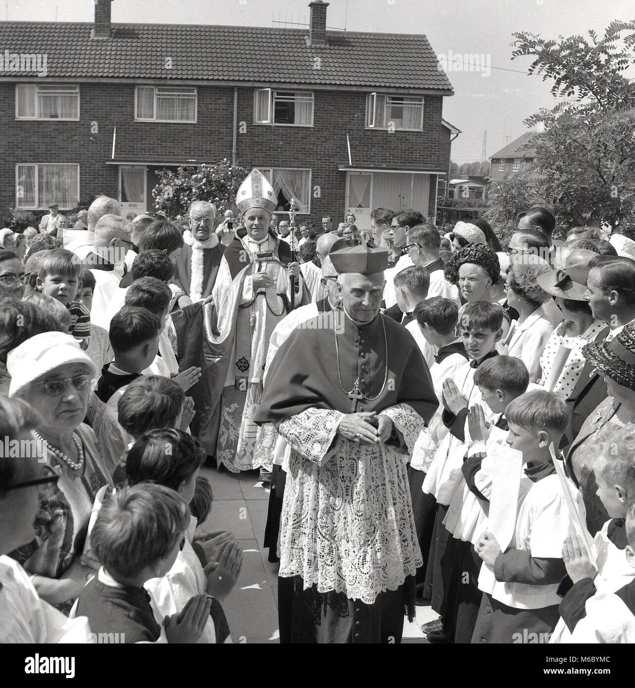 1960, erwachsene Zuschauer und Kinder begrüßen den Besuch von einem römisch-katholischen Bischof zu ihrer Nachbarschaft, ein neuer, moderner 60s style Gehäuse, Bucks, England, UK. Stockfoto