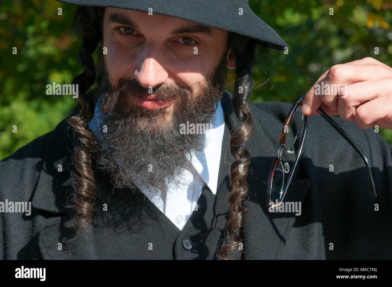 Außenbereich sonnige helle Porträt eines lächelnden Jungen orthodoxen jüdischen Mann mit schwarzen Bart, Hut und Kostüm holding Brillen Stockfoto