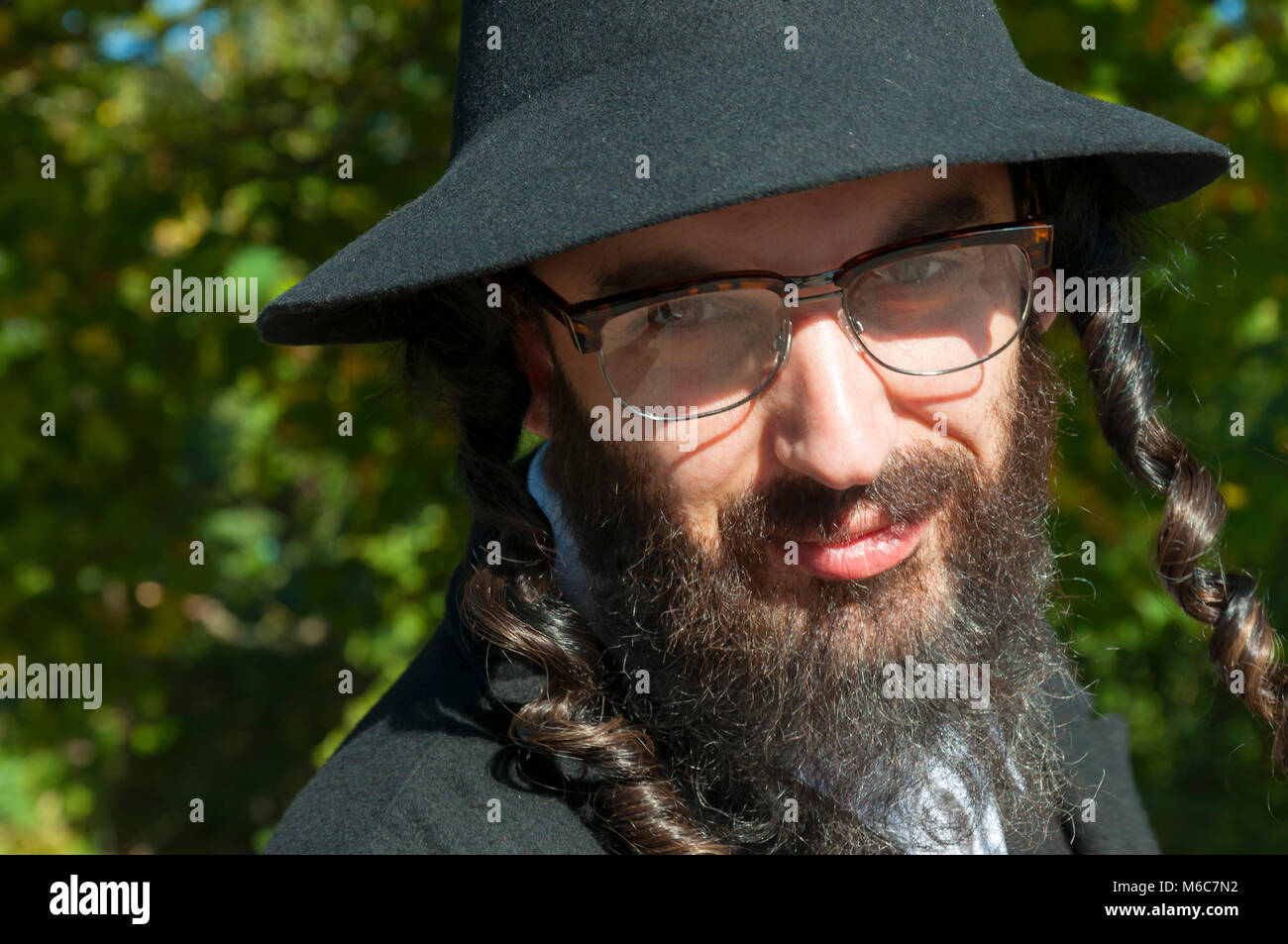 Außenbereich sonnige Porträt eines lächelnden Jungen orthodoxen jüdischen Mann mit schwarzen Bart, Kostüm und Hut Brille tragen Stockfoto