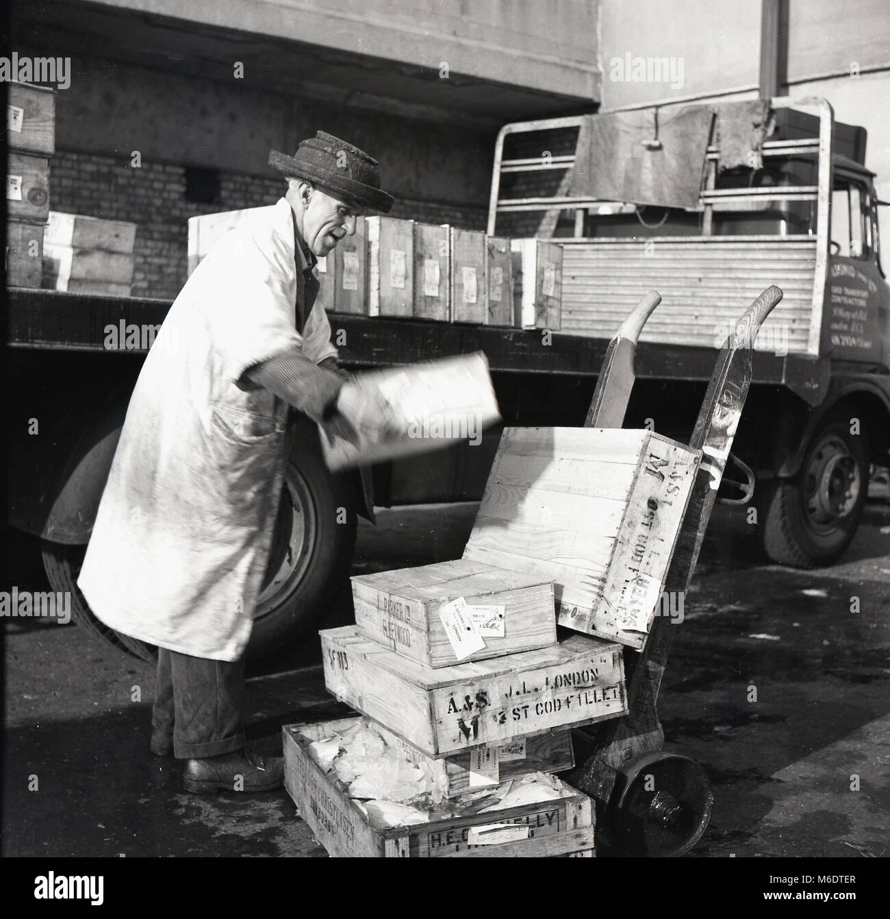 1950er Jahre, historisches Bild eines älteren Pförtners, der Holzkisten aus Kabeljaufilet von einem Lastwagen auf einen Sackkarren- oder Portierwagen vor dem Billingsgate Wholesale Fish Market, London, England, Großbritannien entlädt. Billingsgate, an der Lower Thames Street in der Stadt London, war zu einem Zeitpunkt der größte Fischmarkt der Welt. Stockfoto