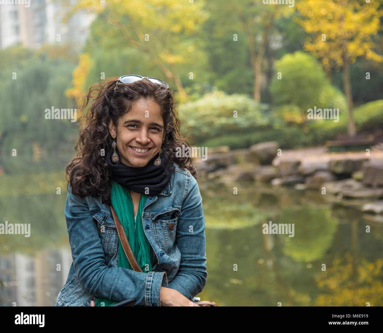 Eine schöne Latina (peruanischen) Frau (Alter 40) lächeln in die Kamera. Sie sitzt vor einem Teich mit Herbstfarben hinter ihr. Sie ist glücklich. Stockfoto