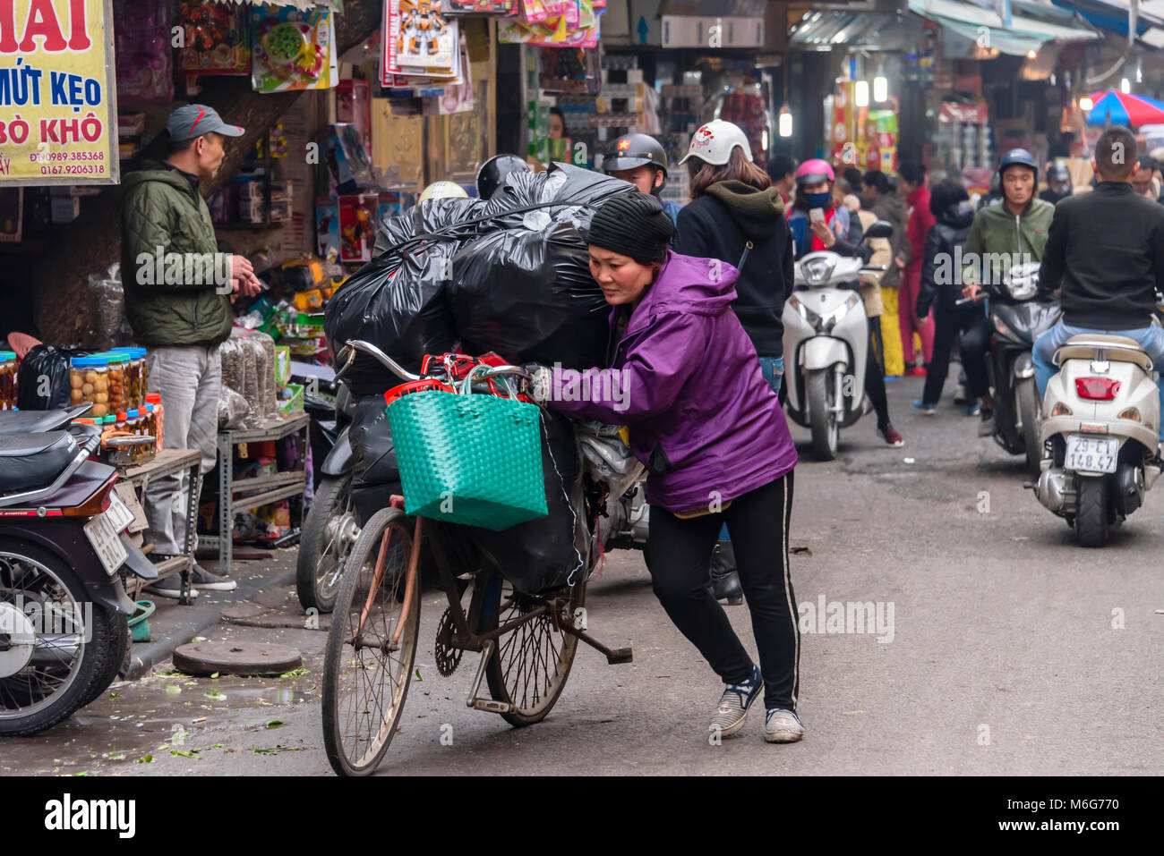 Eine Frau drückt ihr Fahrrad mit schweren schwarzen Beutel in Hanoi, Vietnam geladen Stockfoto