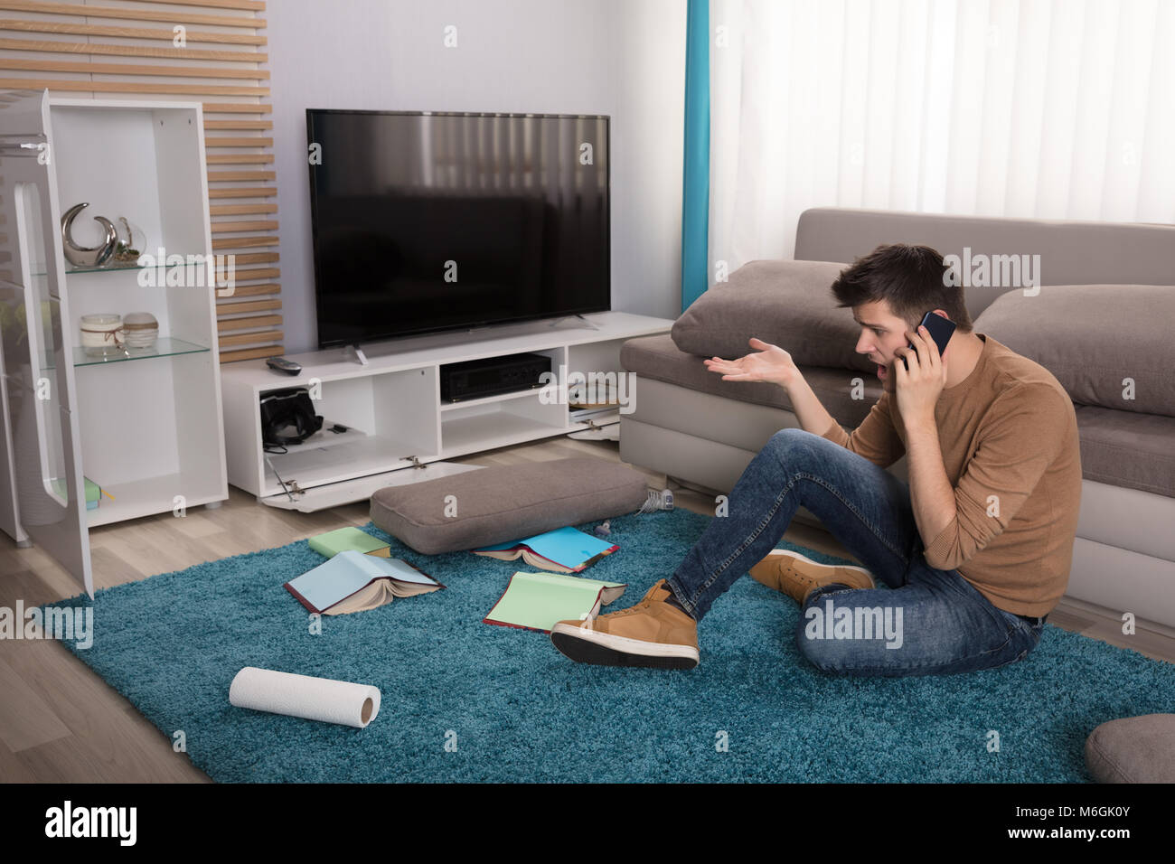 Unglückliche junge Mann sitzt auf Teppich in unordentlichen Zimmer und am Telefon sprechen zu Hause Stockfoto