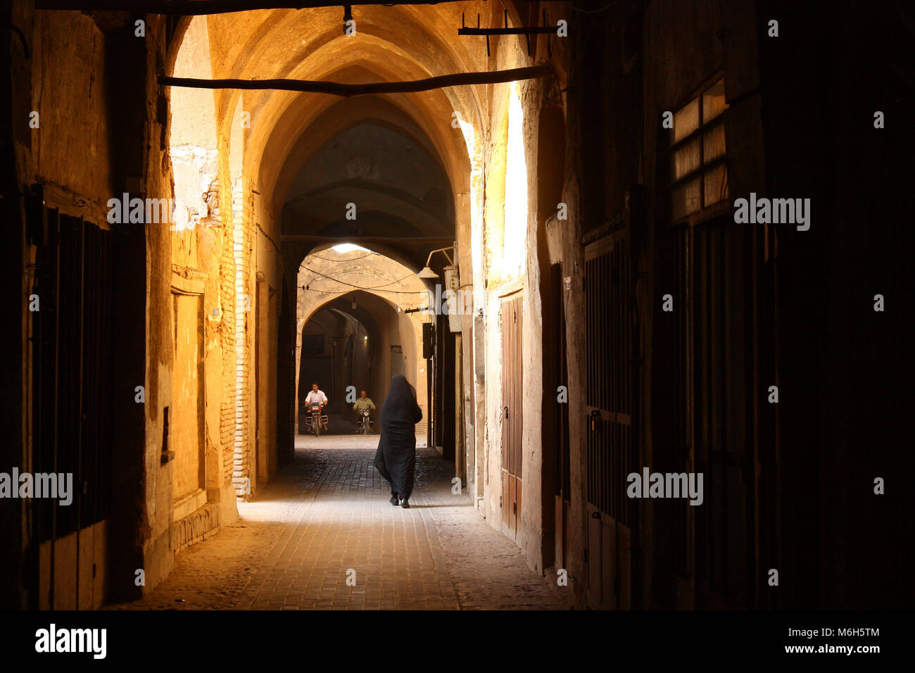 Frau im schwarzen Tschador wandern durch die sonnendurchfluteten historischen adobe Gängen des alten überdachten Basar in Yazd, Iran. 2 Männer auf Motorrädern fahren durch. Stockfoto