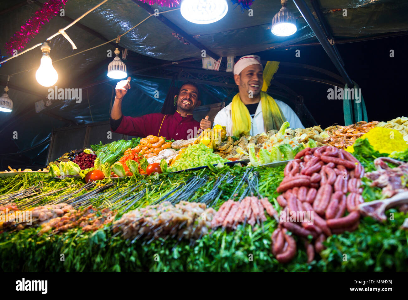 Köche lächelnd an einer Garküche voll mit frischem Gemüse und Fleisch am Spieß in Jemaa el-Fnaa Markt, Marrakesch, Marokko Stockfoto