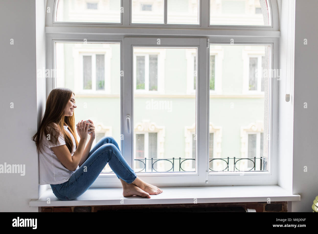 Schöne Frau Kaffee trinken und Blick durch Fenster beim Sitzen am Fensterbrett zu Hause Stockfoto
