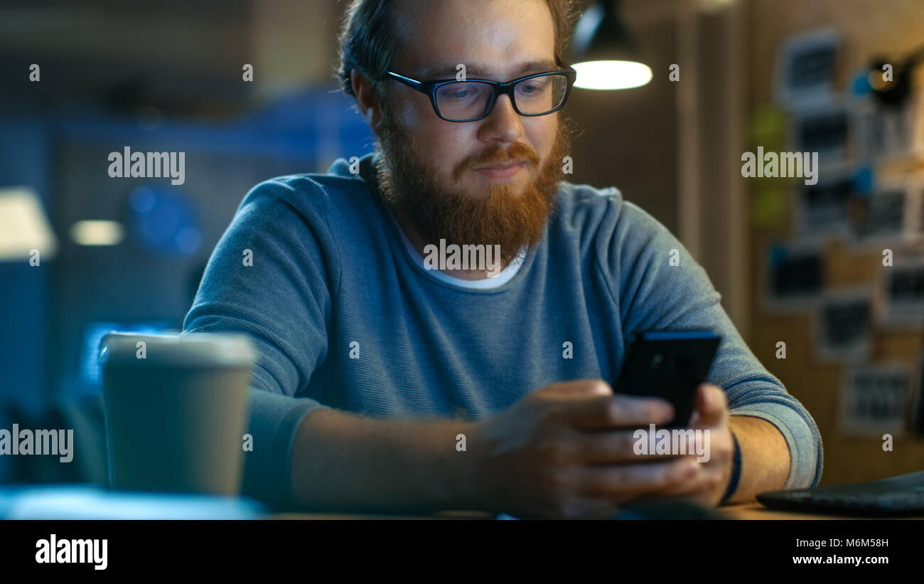 Junger bärtiger Mann Typen Nachricht auf seinem Smartphone. Er sitzt am Schreibtisch und lächelt. Stockfoto