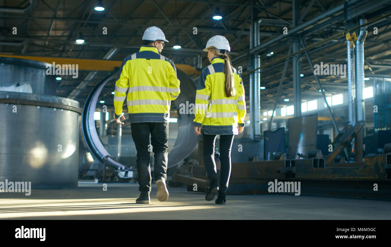 Ansicht von hinten erschossen von Männlichen und Weiblichen Wirtschaftsingenieure in Diskussion während man durch Heavy Industry Manufacturing Factory. Große Metallarbeiten Stockfoto